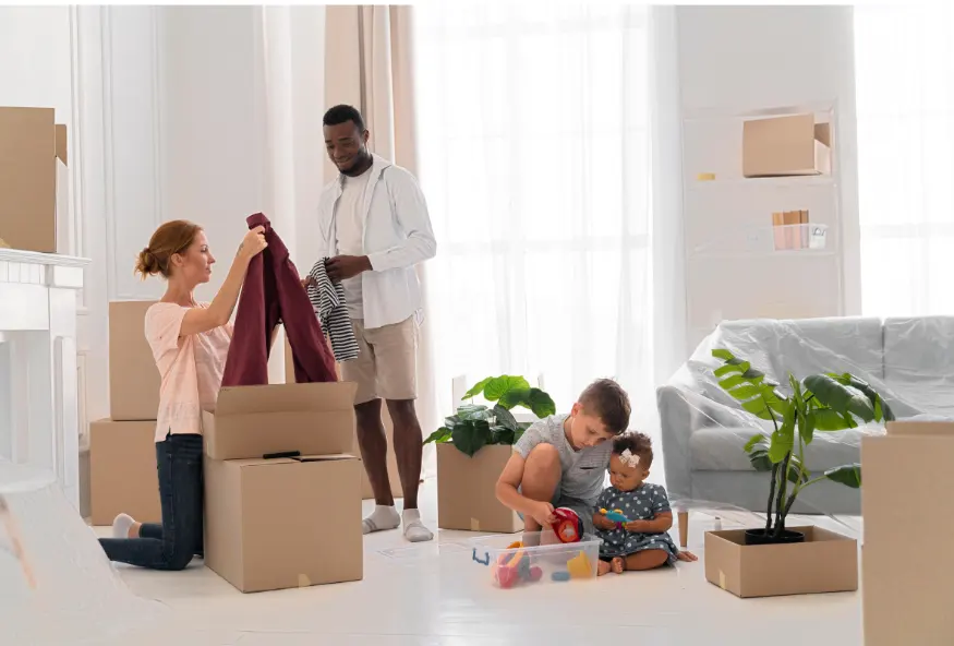 In the image, a family of four is seen in a bright room, possibly in the middle of moving. On the left, a red-haired woman, dressed in a short-sleeved shirt and jeans, is kneeling next to an open box, either taking out or putting away clothes. Near her, a man standing, dressed in a white shirt and beige shorts, is also handling clothes. To the right, a young boy, sitting on the floor, is playing with toys next to a baby. The boy is wearing a gray t-shirt and the baby a polka-dotted dress. The room is filled with moving boxes and some potted plants. In the background, there is a sofa covered with a sheet and partially filled shelves.