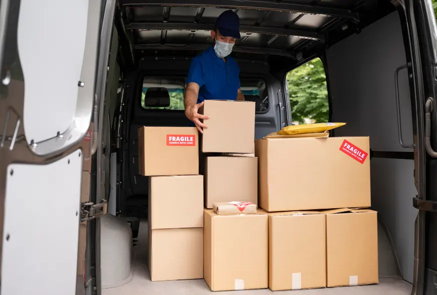 In the image, a man is seen inside a cargo van, organizing cardboard boxes. The man, wearing a blue cap, a blue shirt, and a face mask, is carefully placing a box on top of a stack of other boxes. Some of the boxes have red labels indicating they are fragile. The van is parked with the rear door open, allowing a view of the partially filled interior. The scene suggests that the man is in the middle of a delivery or moving process.