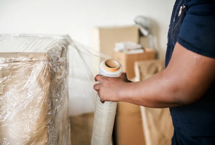 A close-up of a person wrapping a large object with clear plastic. Boxes can be seen in the background, suggesting that the person is in the process of packing for a move or storage.