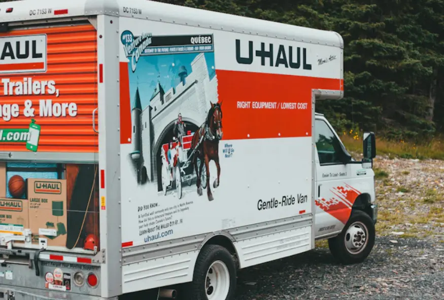 A moving truck from the company U-Haul, with a Quebec design on the side. The truck is parked outdoors, next to a vegetated area.